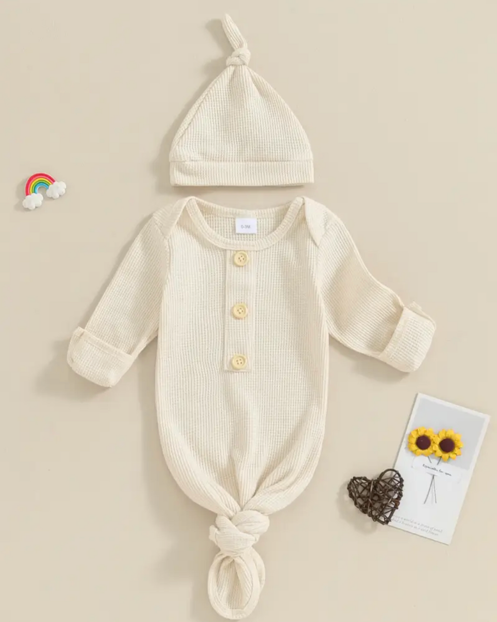 A baby knit three-piece outfit set featuring a long-sleeve romper with a front button closure, a matching knotted bonnet, and a small card with sunflowers, arranged on a beige background.
