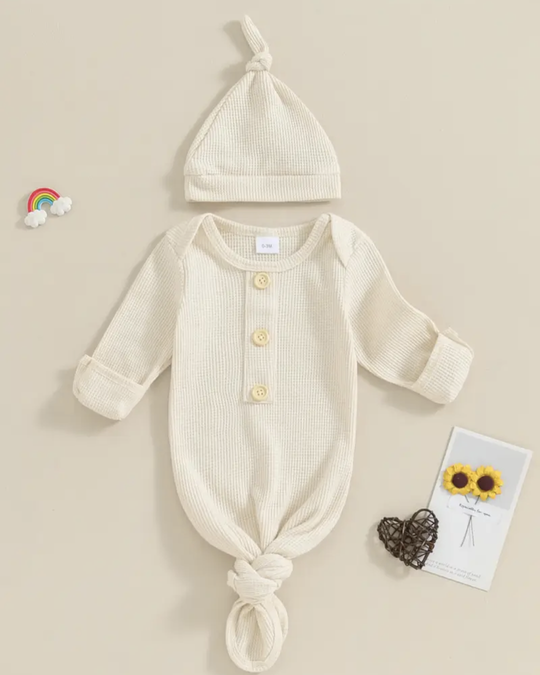 A baby knit three-piece outfit set featuring a long-sleeve romper with a front button closure, a matching knotted bonnet, and a small card with sunflowers, arranged on a beige background.