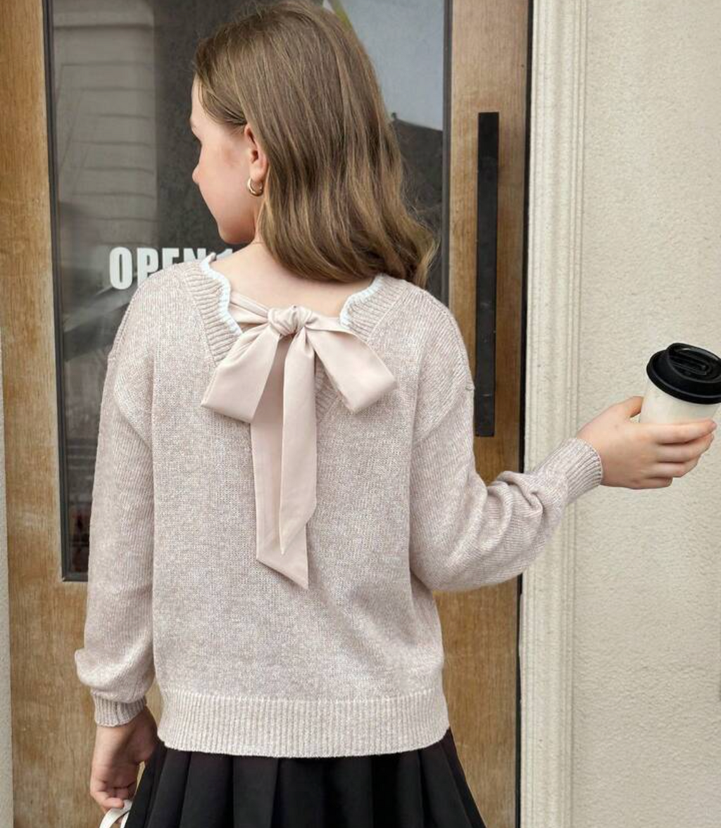 A girl viewed from behind wearing a light beige knit sweater with a large bow detail on the back, paired with a black skirt. She is holding a takeaway coffee cup next to a doorway.