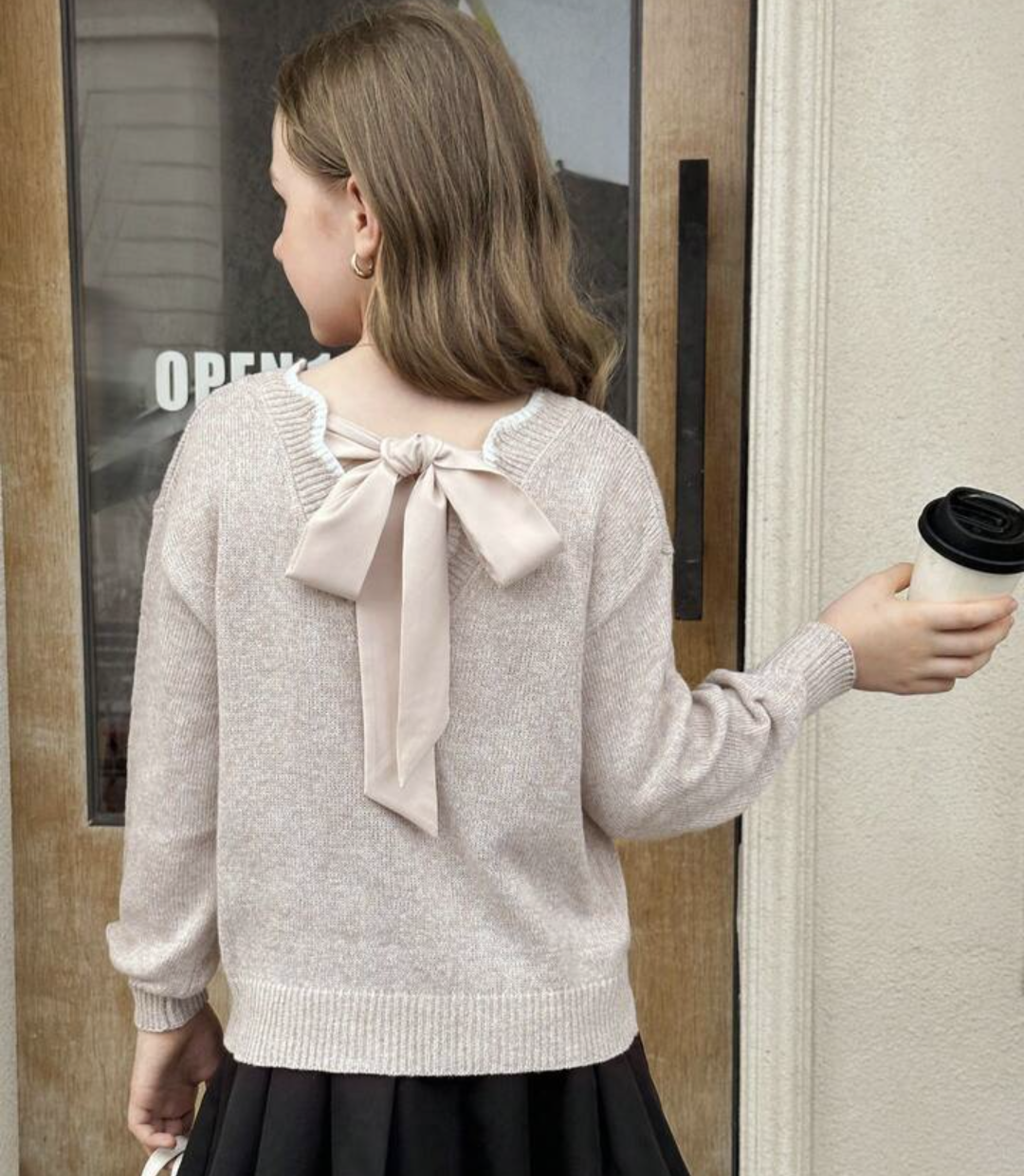 A girl viewed from behind wearing a light beige knit sweater with a large bow detail on the back, paired with a black skirt. She is holding a takeaway coffee cup next to a doorway.