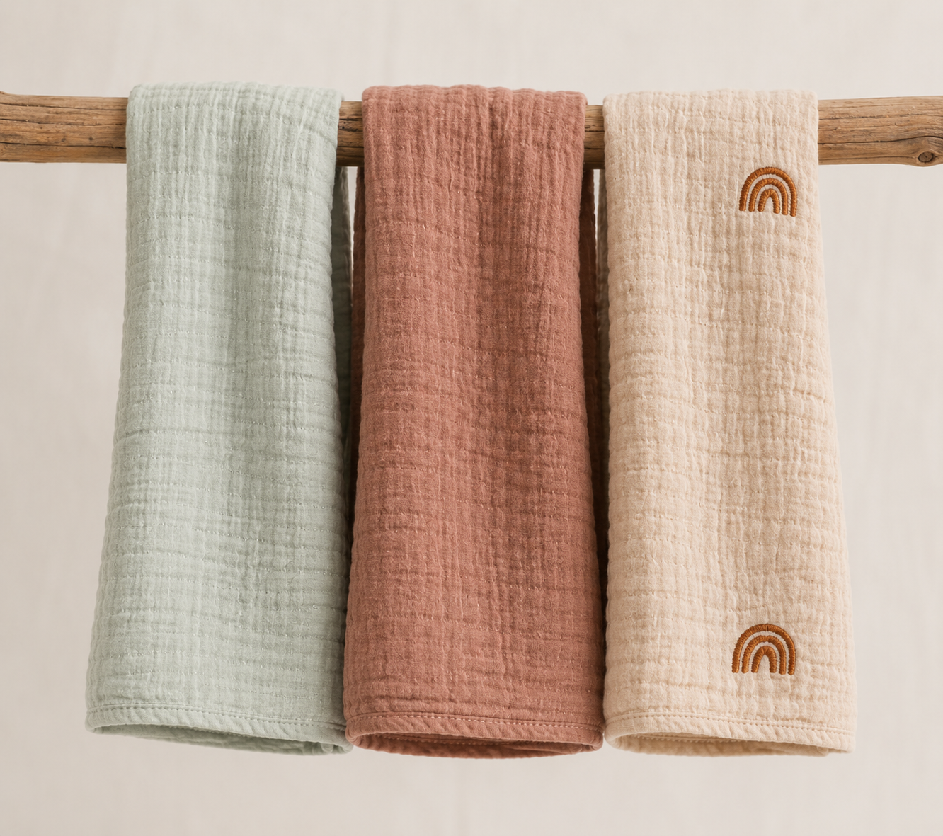 Three textured cotton muslin towels in sage green, dusty rose, and cream beige hang from a wooden dowel against a light beige background. The cream towel on the right features an embroidered brown rainbow motif.